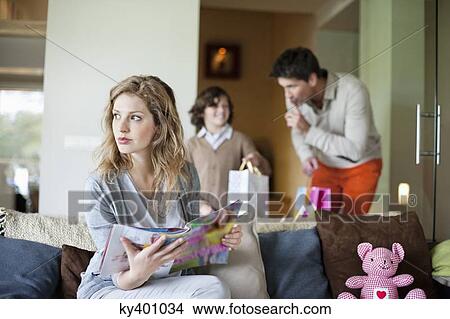 Picture - Woman sitting on couch with her husband and son holding surprise gifts in background at mother day. Fotosearch