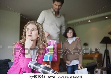 Stock Photo - Woman thinking on couch with her husband and son holding surprise gifts in background at mother day. Fotosearch