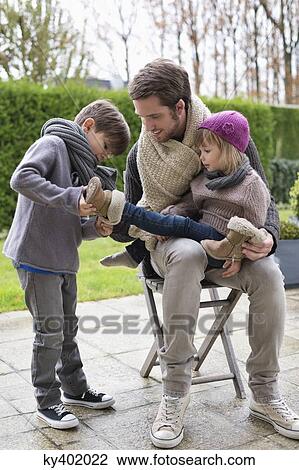 Stock Image - Boy putting shoe on his sister sitting in her father's lap. Fotosearch