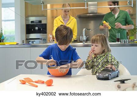 Stock Image - Little girl with a toy car looking at his brother cooking in the kitchen. Fotosearch