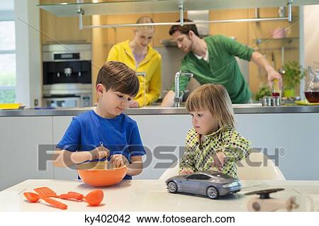 Stock Image - Little girl with a toy car looking at his brother cooking in the kitchen. Fotosearch