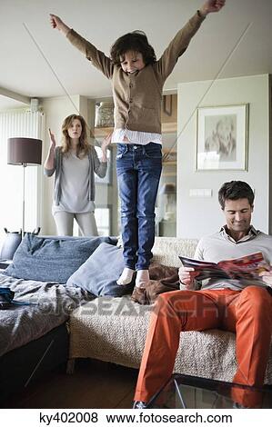 Stock Photo - Naughty boy jumping on couch with his parents at home. Fotosearch