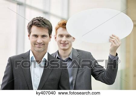 Stock Image - Portrait of a businesswoman smiling with her colleague holding speech bubble behind her. Fotosearch