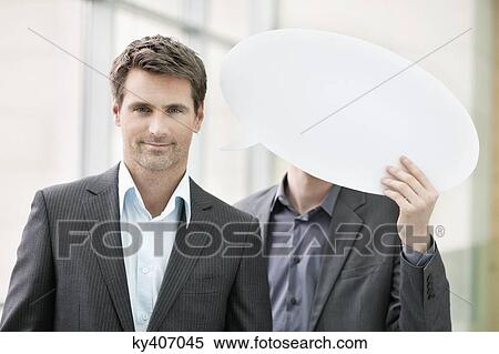 Stock Photography - Portrait of a businesswoman smiling with her colleague holding speech bubble behind her. Fotosearch