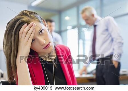 Stock Image - Female executive looking sad in an office with her colleagues discussing in the background. Fotosearch