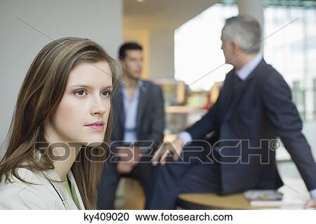 Stock Image - Female executive thinking in an office with her colleagues discussing in the background. Fotosearch