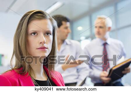 Stock Image - Female executive thinking in an office with her colleagues discussing in the background. Fotosearch