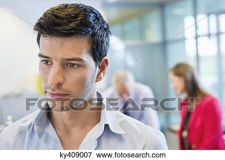 Male executive thinking in an office with his colleagues discussing in the background View Large Photo Image Stock Photo - Male executive thinking in an office with his colleagues discussing in the background. Fotosearch
