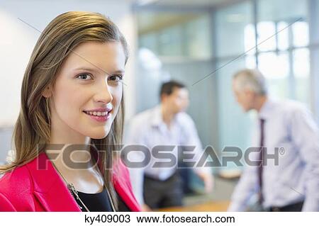 Stock Image - Portrait of a businesswoman smiling in an office with her colleagues discussing in the background. Fotosearch
