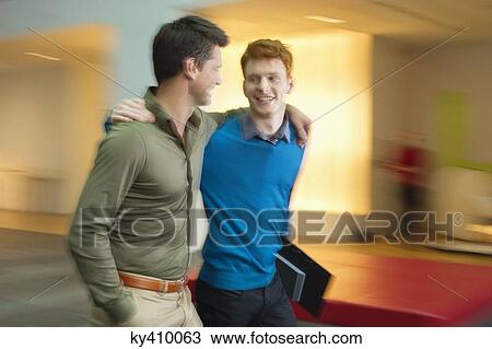 Stock Image - Businessmen walking with their arm around each other in an office corridor. Fotosearch
