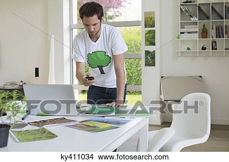 Picture - Man holding a mobile phone and working on a laptop. Fotosearch