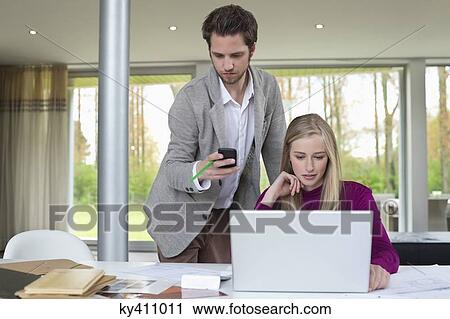 Woman working on a laptop with a man using mobile phone beside her View Large Photo Image Stock Image - Woman working on a laptop with a man using mobile phone beside her. Fotosearch