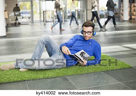 Businessman listening to music and reading book while relaxing on grass mat in an office lobby View Large Photo Image Stock Image - Businessman listening to music and reading book while relaxing on grass mat in an office lobby. Fotosearch