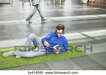 Businessman listening to music and reading book while relaxing on grass mat in an office lobby View Large Photo Image Stock Photography - Businessman listening to music and reading book while relaxing on grass mat in an office lobby. Fotosearch
