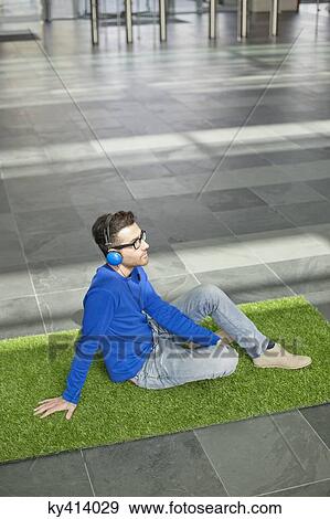 Businessman relaxing on grass and listening to music in an office lobby View Large Photo Image Stock Photo - Businessman relaxing on grass and listening to music in an office lobby. Fotosearch