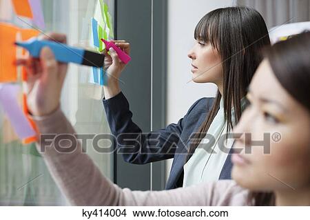 Businesswomen writing on adhesive notes in an office View Large Photo Image Picture - Businesswomen writing on adhesive notes in an office. Fotosearch
