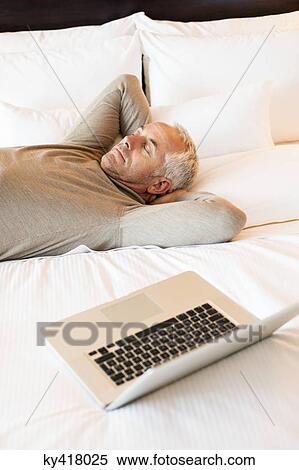 Stock Photography - Man resting on the bed with a laptop in a hotel room. Fotosearch