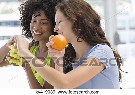 Stock Photo - Close-up of two smiling female friends eating fruits. Fotosearch