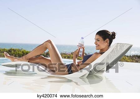 Picture - Beautiful woman holding a water bottle while sunbathing on the beach. Fotosearch