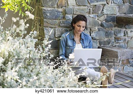 Stock Image - Woman leaning against a stone wall and using a laptop. Fotosearch