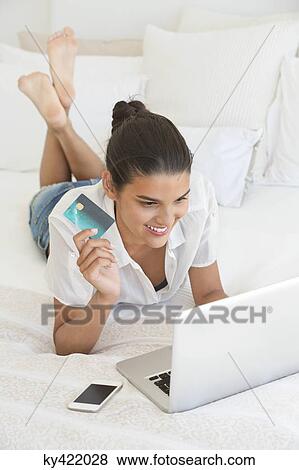 Woman using laptop and holding a credit card on the bed View Large Photo Image Stock Photo - Woman using laptop and holding a credit card on the bed. Fotosearch