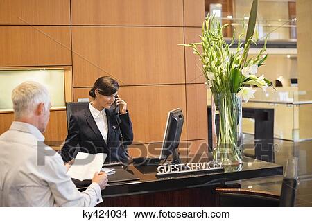 Businessman reading a brochure with a receptionist talking on a telephone at the hotel reception counter View Large Photo Image Picture - Businessman reading a brochure with a receptionist talking on a telephone at the hotel reception counter. Fotosearch
