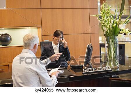 Stock Photography - Receptionist talking on a landline phone with a businessman pointing at the hotel reception counter. Fotosearch