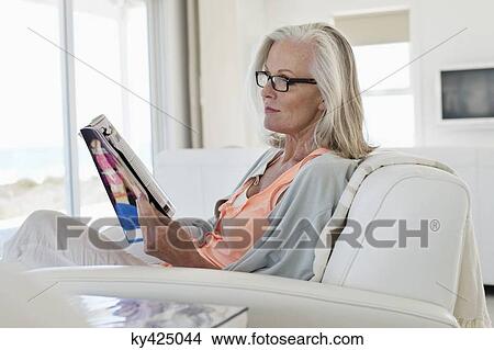 Picture - Woman sitting on a couch and reading a magazine at home. Fotosearch