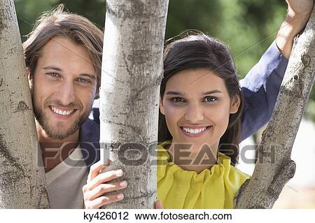 Stock Image - Portrait of a couple smiling behind a tree. Fotosearch