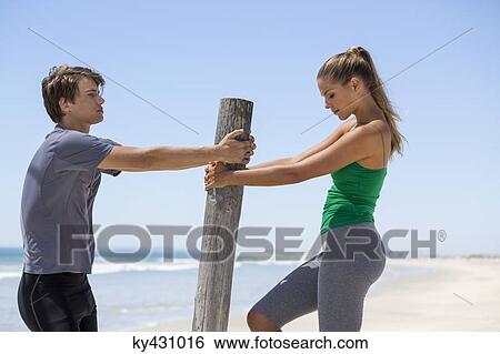 Stock Photograph - Man assisting a woman to exercise on the beach. Fotosearch