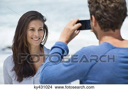 Man taking a picture of his wife with a cell phone on the beach View Large Photo Image Picture - Man taking a picture of his wife with a cell phone on the beach. Fotosearch