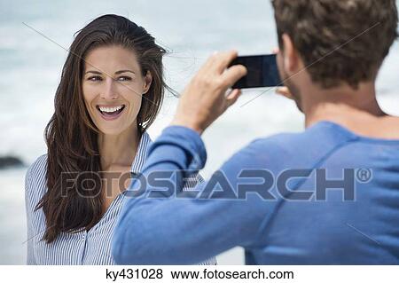 Man taking a picture of his wife with a cell phone on the beach View Large Photo Image Stock Photo - Man taking a picture of his wife with a cell phone on the beach. Fotosearch