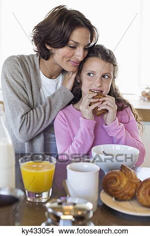 Picture - Girl having breakfast beside her mother at a kitchen counter. Fotosearch