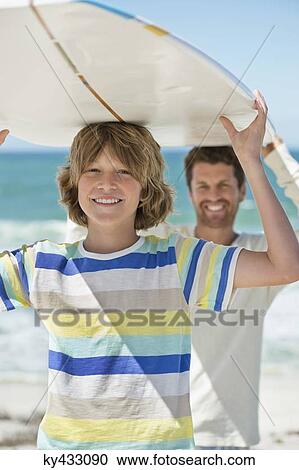 Stock Image - Man and his son carrying a surfboard on the beach. Fotosearch
