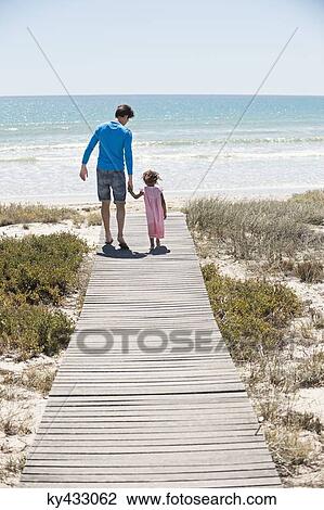 Stock Image - Man with his daughter walking on a boardwalk on the beach. Fotosearch