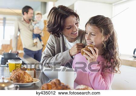 Stock Image - Girl having breakfast beside her mother at a kitchen counter. Fotosearch