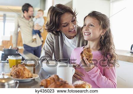 Stock Image - Girl having breakfast beside her mother at a kitchen counter. Fotosearch