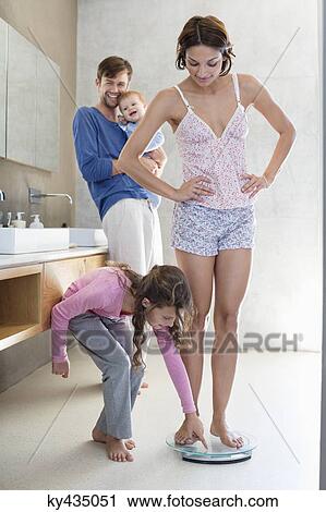 Woman measuring her weight on a weighing scale with her family in bathroom View Large Photo Image Stock Image - Woman measuring her weight on a weighing scale with her family in bathroom. Fotosearch