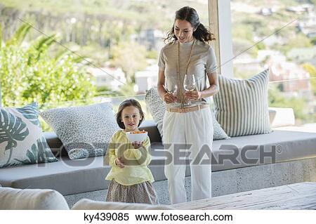 Stock Photography - Girl holding a bowl of snacks beside her mother holding stem glasses at home. Fotosearch