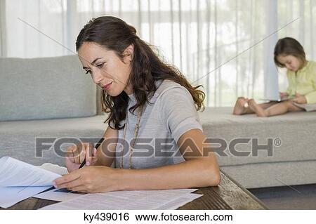Stock Photograph - Woman doing paperwork with her daughter using a digital tablet in the background. Fotosearch