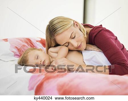 Picture - Woman resting her head on her baby hand sleeping on the bed. Fotosearch