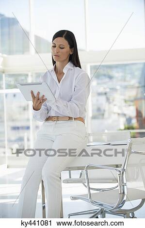 Businesswoman using a digital tablet while sitting on a table in an office View Large Photo Image Stock Image - Businesswoman using a digital tablet while sitting on a table in an office. Fotosearch