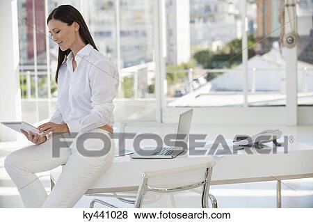 Businesswoman using a digital tablet while sitting on a table in an office View Large Photo Image Stock Photo - Businesswoman using a digital tablet while sitting on a table in an office. Fotosearch