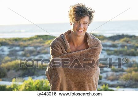 Stock Photograph - Happy man wrapped in a towel standing on the beach. Fotosearch