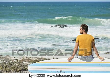 Stock Image - Man sitting on a surfboard on the beach. Fotosearch