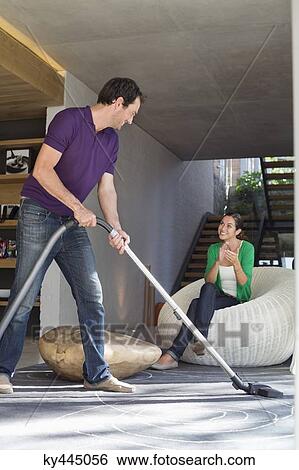 Stock Photograph - Man cleaning house with a vacuum cleaner with his wife sitting on a seat. Fotosearch