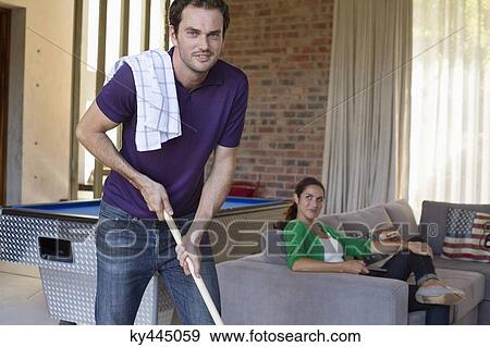 Stock Photo - Man holding a mop with his wife sitting on a couch. Fotosearch