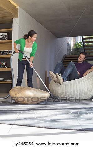 Woman cleaning house with a vacuum cleaner with her husband sitting on a seat View Large Photo Image Picture - Woman cleaning house with a vacuum cleaner with her husband sitting on a seat. Fotosearch