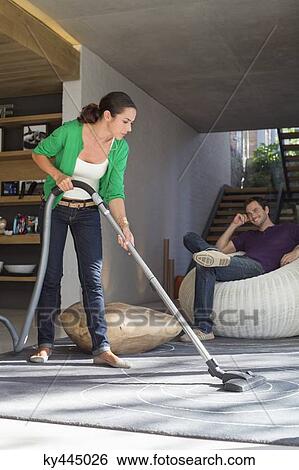 Stock Photograph - Woman cleaning house with a vacuum cleaner with her husband sitting on a seat. Fotosearch