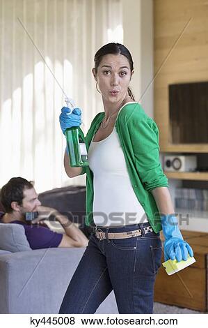 Stock Photo - Woman holding cleaning equipment with her husband sitting on a couch. Fotosearch